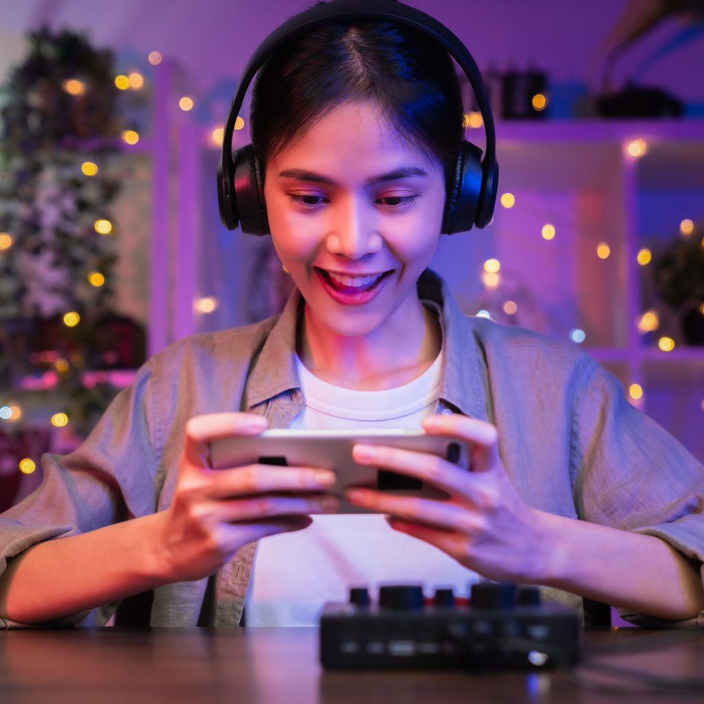 Excited Young Asian woman wearing headset and playing online game on smartphone with live broadcasting on internet.