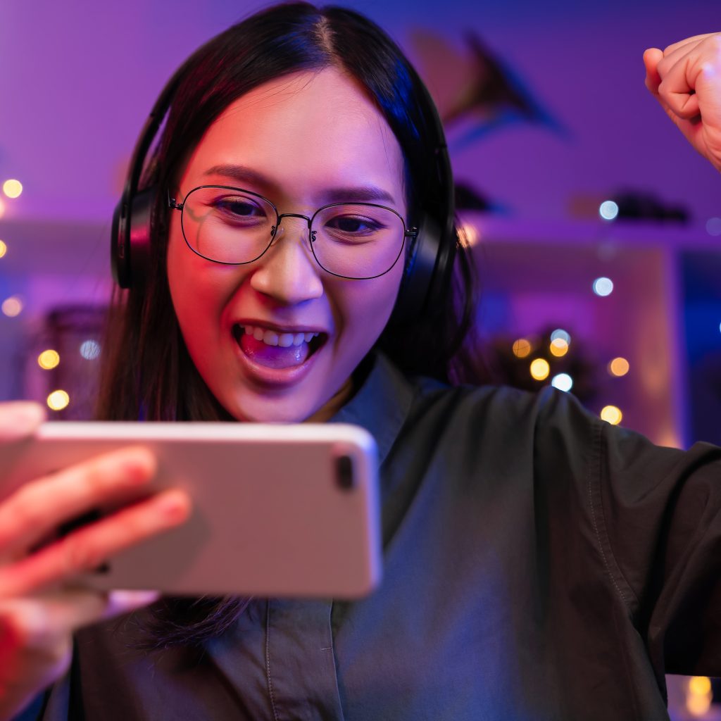 Excited Young Asian woman wearing headset and playing online game on smartphone with live broadcasting on internet.