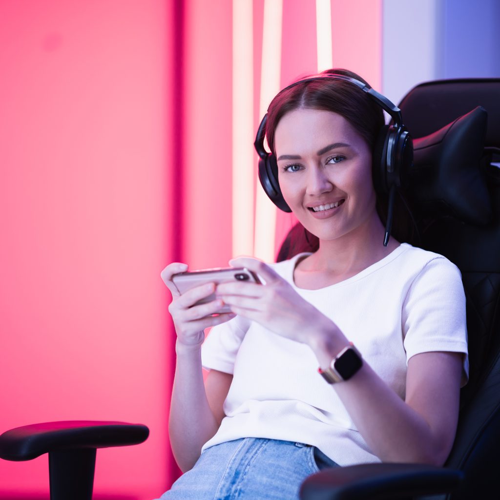 Gamer girl in a big professional music headphones in white t-shirt and blue jeans holds smart phone in hands and plays game app on mobile phone, smiling and looking at camera indoors.