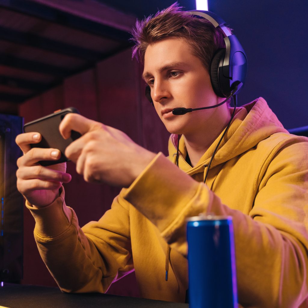 Portrait of young guy pro gamer wearing headset streaming while playing mobile video game on his smartphone in neon coloured room at home. Focused streamer participates in online e-sport tournament