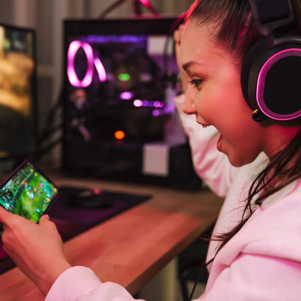 Young happy woman gamer playing video games on smartphone and computer in dark room wearing headphones