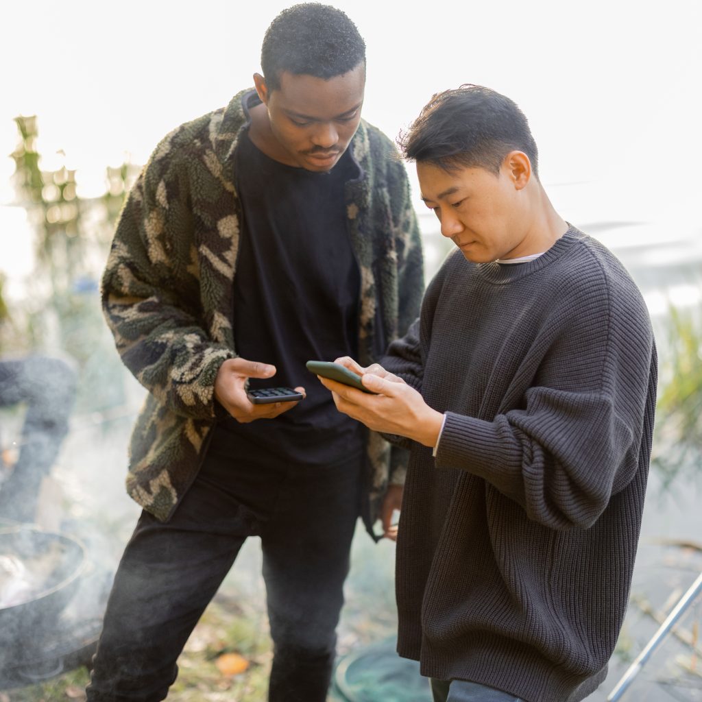 Asian and latin men using smartphones during resting on river or pond coast. Men fishing and cooking on river coast at autumn. Leisure and weekend in nature. Idea of friendship and spend time together