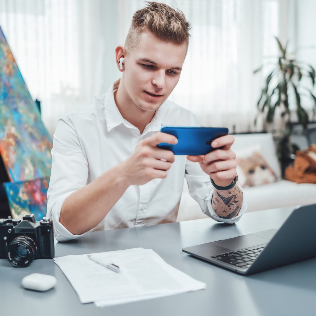 Male designer with photo camera and laptop doing his job in modern office. Tattooed guy dressed in white shirt sits at table using his phone.