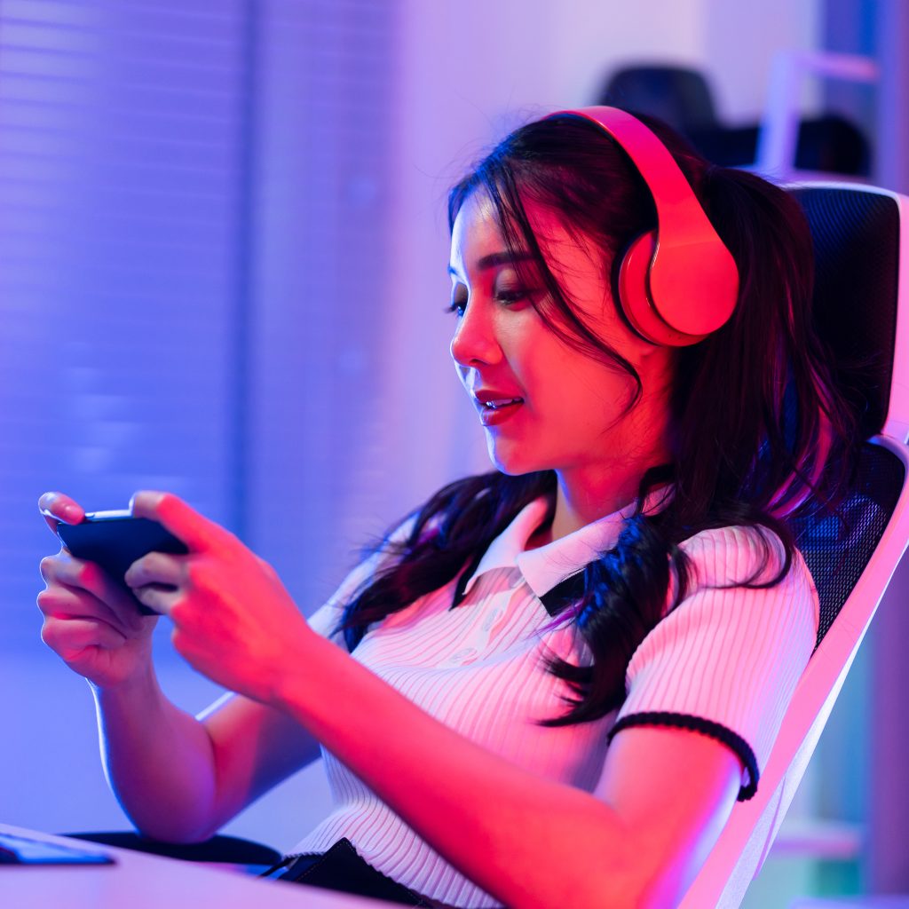 Asian young beautiful woman playing mobile game on smartphone at home. Attractive casual girl feel happy and relax sit on chair having fun touching screen on phone to play video enjoy victory in house