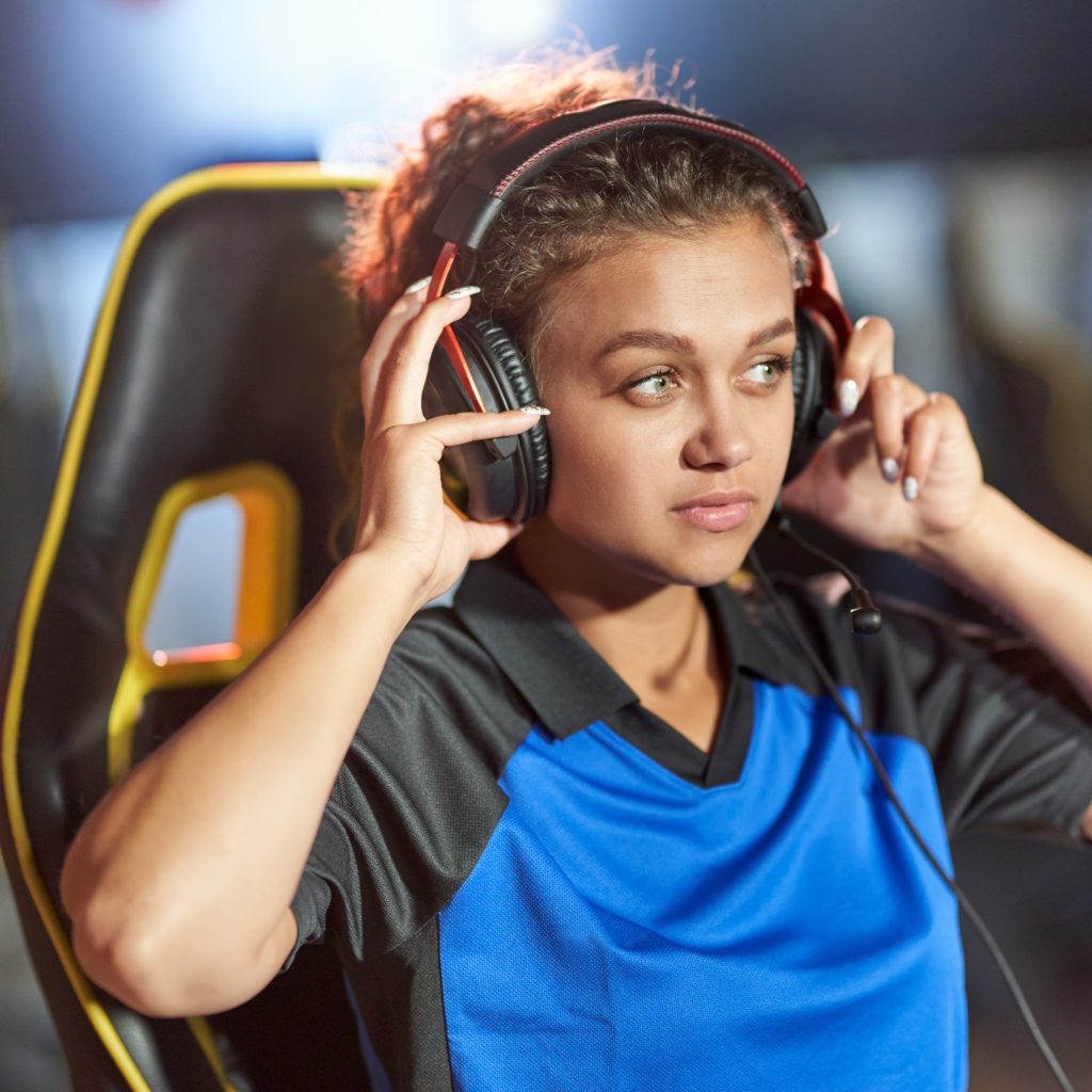 Professional cyber sport. Teenage mixed race girl wearing headphones playing online video games, sitting on chair and looking at PC screen. eSports Tournament concept