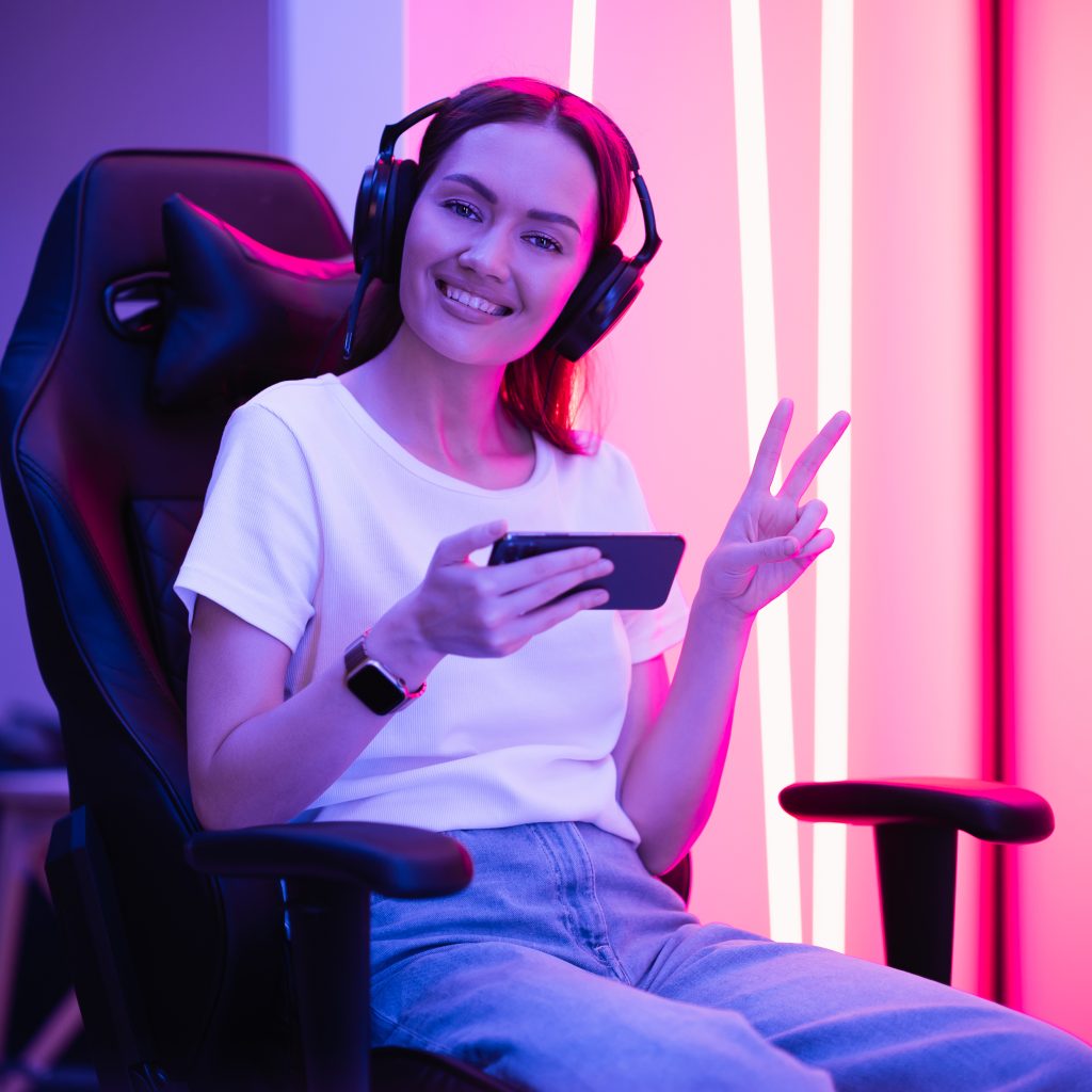 Gamer girl in white t-shirt and blue jeans holds smart phone in hands and plays game app on mobile phone, smiling and looking at cellphone screen indoors