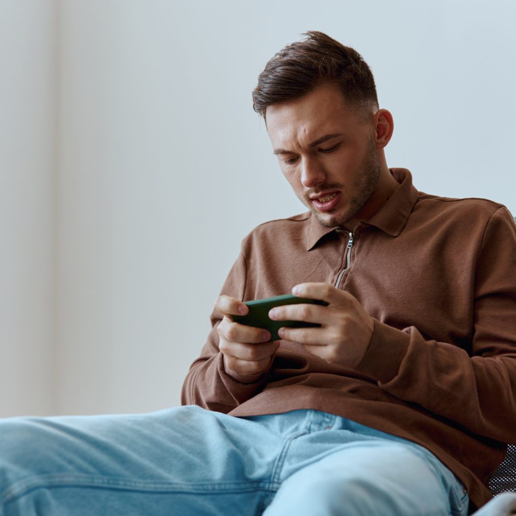 Mobile App concept. Concentrated focused confused attractive try to win playing in exciting raising game opening mouth looks at screen. Happy guy relaxing with phone chatting with friends. Copy space
