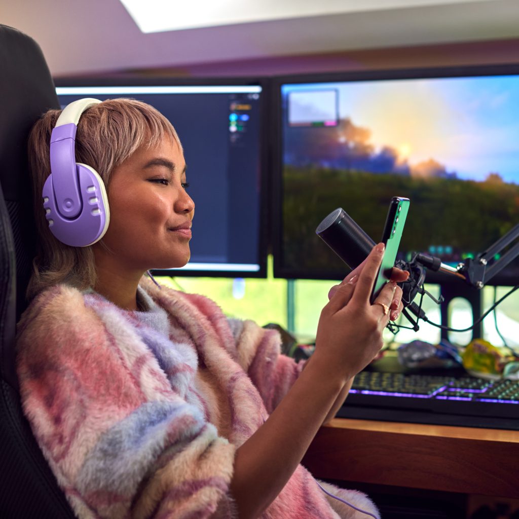 Woman With Mobile Phone Gaming At Home Sitting At Desk With Multiple Monitors