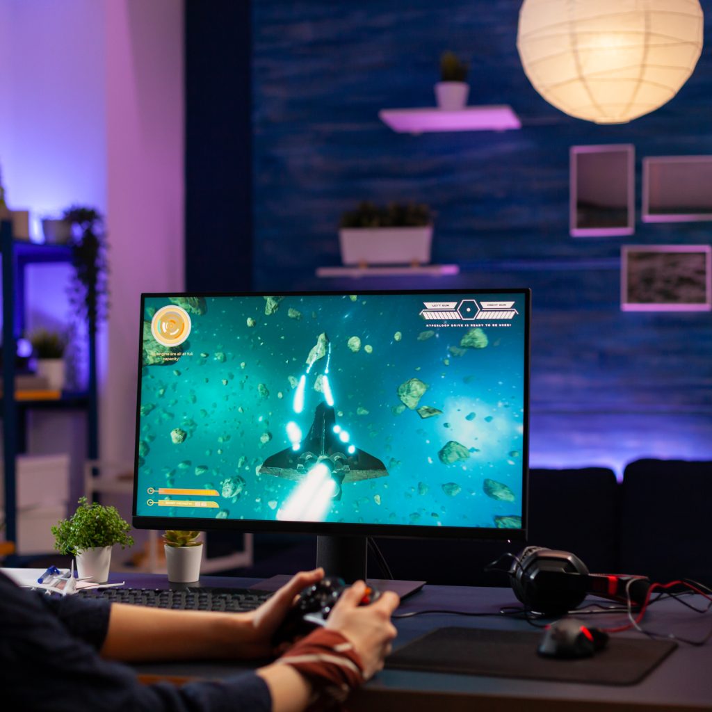 Concentrated player sitting on gaming desk late at night using wireless controller playing space shooter. Virtual streaming cyber performing live tournament using professional equipment