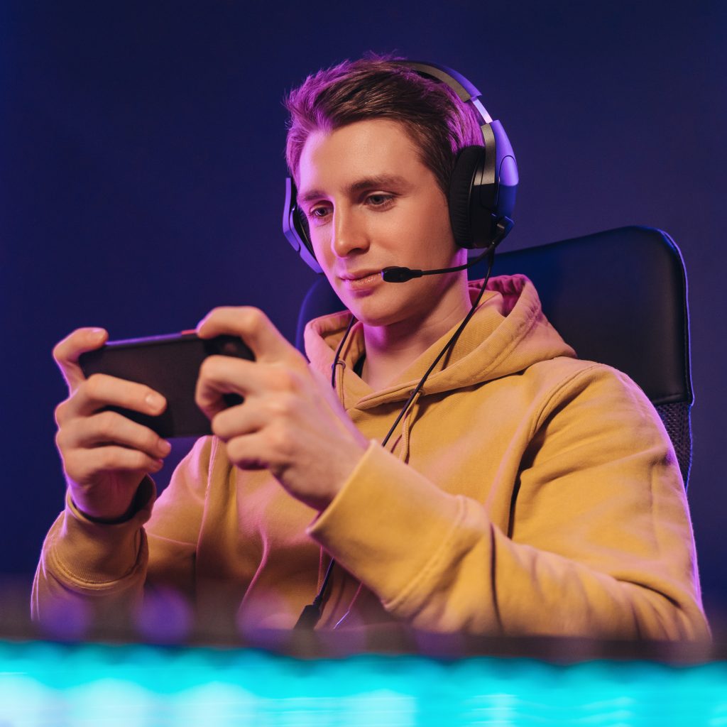 Portrait of caucasian man pro gamer sitting on gaming chair, wearing headset, has live stream while playing mobile game on the smartphone at home. Young cyber sportsman holding cellphone in hands