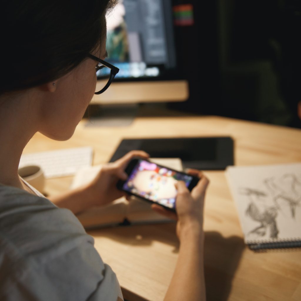 Cropped image of young lady designer sitting indoors at night play games by mobile phone.
